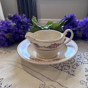 May include: A cream-colored gravy boat and saucer set with a delicate floral pattern and gold trim. The set is placed on a white lace tablecloth, with purple flowers in the background. The gravy boat has a handle and a spout.