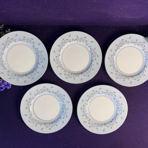 May include: A set of five round white ceramic plates featuring a delicate light blue floral vine pattern along the rim. The plates have a smooth, solid white centre and are arranged in a pentagonal formation against a deep purple background.