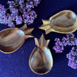 May include: Three wooden pineapple-shaped bowls with a natural wood grain finish are arranged on a dark blue surface. Purple lilac flowers are placed around the bowls. The bowls have a carved design and a shallow interior.