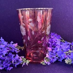May include: A translucent magenta glass vase featuring an etched floral pattern of sunflowers and delicate leaves. The vase has a scalloped rim and a tapered cylindrical shape, surrounded by clusters of vibrant purple flowers at the base.