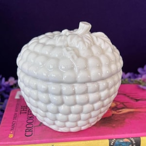May include: A white ceramic lidded jar shaped like a cluster of raspberries. The surface features a repeating embossed spherical texture mimicking individual fruit drupelets, topped with a small sculpted stem and leaf detail on the lid.