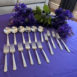 May include: A collection of silver cutlery, including spoons and forks, displayed on a purple tablecloth. The cutlery features a floral design on the handles. A bunch of purple flowers is arranged in the background.