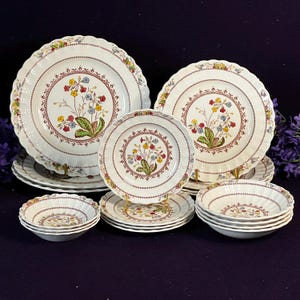 May include: A collection of cream-colored ceramic dinnerware featuring scalloped edges and a central botanical motif of red, yellow, and blue flowers with green leaves. The set includes various sizes of plates and bowls with a textured rim pattern.