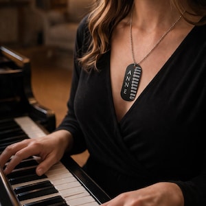 May include: A black dog tag necklace with the name "ANNE" vertically printed in white. The tag has a silver chain and is worn over a black top. The image shows a person playing a piano.