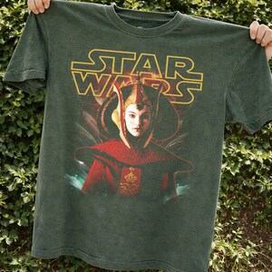 Puede incluir: Camiseta verde oscuro con el logo dorado de Star Wars. La camiseta muestra un retrato de un personaje con ropa roja y dorada. La imagen es un art&iacute;culo promocional de la franquicia Star Wars.