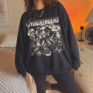 以下が含まれることがあります： ダークチャコールグレーのクルーネックトレーナー。スーパーヒーローのポーズをとった様々な漫画キャラクターのグラフィックプリントが施されています。キャラクターの上にはかすれた白文字で「MICKENGERS」とプリントされ、下部には「MARVEL」の文字があります。