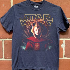 Puede incluir: Camiseta azul marino de Star Wars con un gr&aacute;fico de Padm&eacute; Amidala con su atuendo real. El logotipo dorado de Star Wars est&aacute; encima de la imagen. La camiseta est&aacute; colgada de una percha met&aacute;lica contra una pared de ladrillos.