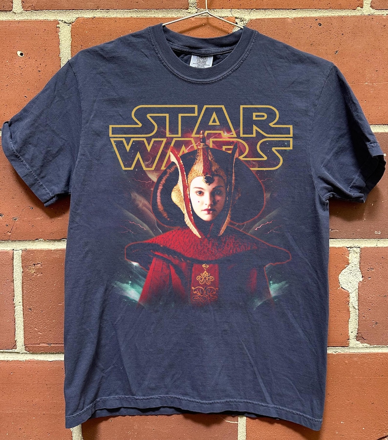 Puede incluir: Camiseta azul marino de Star Wars con un gr&aacute;fico de Padm&eacute; Amidala con su atuendo real. El logotipo dorado de Star Wars est&aacute; encima de la imagen. La camiseta est&aacute; colgada de una percha met&aacute;lica contra una pared de ladrillos.