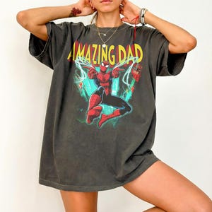 May include: Dark gray t-shirt featuring a graphic of Spider-Man with the text "AMAZING DAD" in yellow and red. The superhero is depicted in a dynamic pose with web-like lines. The shirt has a vintage, worn look.