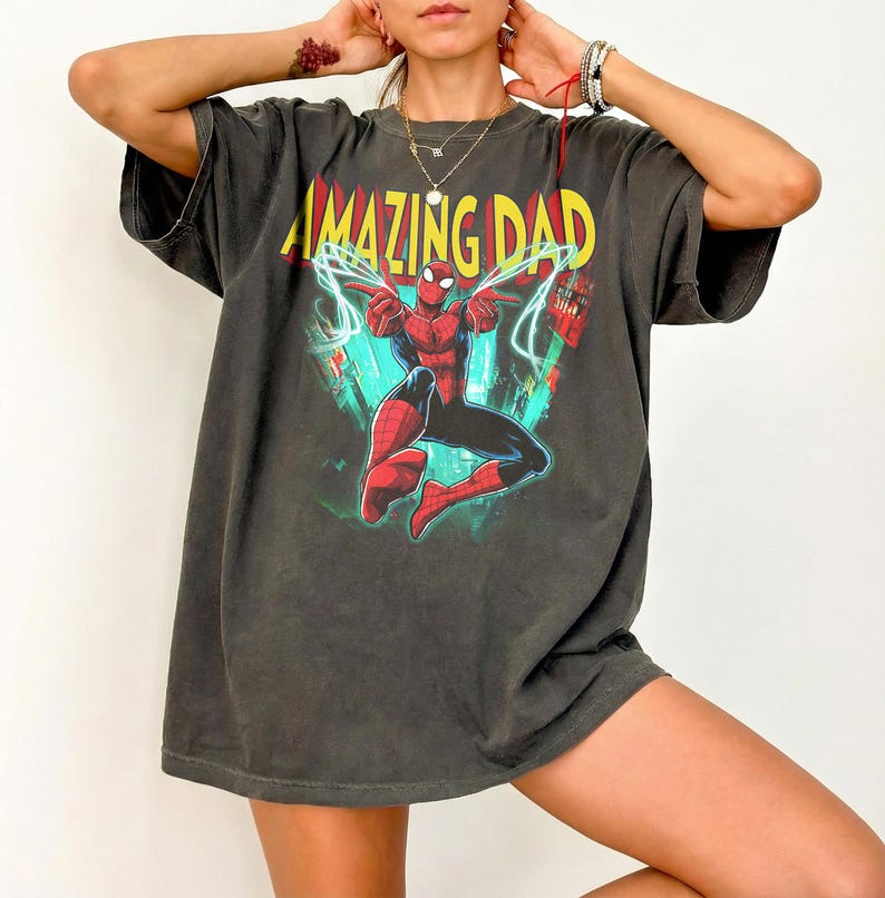 May include: Dark gray t-shirt featuring a graphic of Spider-Man with the text "AMAZING DAD" in yellow and red. The superhero is depicted in a dynamic pose with web-like lines. The shirt has a vintage, worn look.