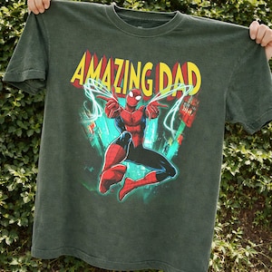 May include: Dark green t-shirt featuring a graphic of Spider-Man with the text "AMAZING DAD" in yellow and red. The superhero is in a dynamic pose with web-like lines. The shirt is held up against a green leafy background.
