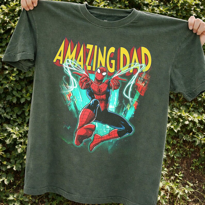 May include: Dark green t-shirt featuring a graphic of Spider-Man with the text "AMAZING DAD" in yellow and red. The superhero is in a dynamic pose with web-like lines. The shirt is held up against a green leafy background.