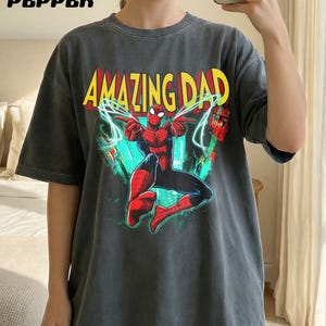 May include: Dark gray t-shirt featuring the text "AMAZING DAD" in yellow and red, with a graphic of Spider-Man. The superhero is depicted in his classic red and blue suit, with web lines and cityscapes in the background.