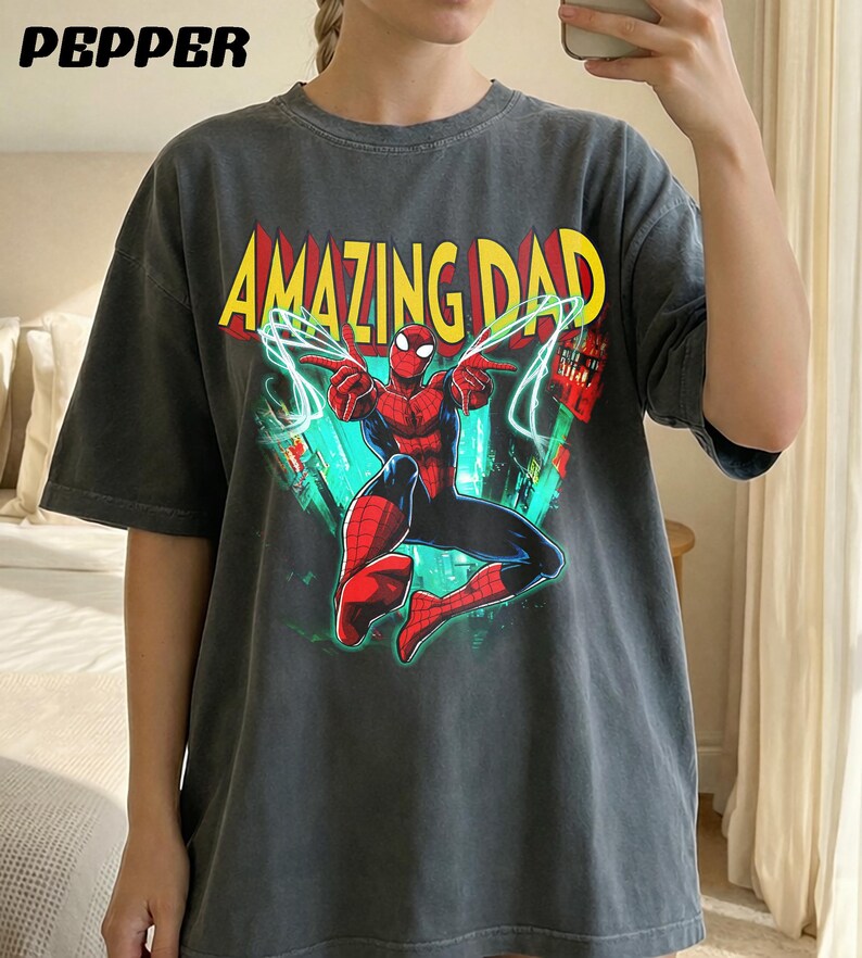 May include: Dark gray t-shirt featuring the text "AMAZING DAD" in yellow and red, with a graphic of Spider-Man. The superhero is depicted in his classic red and blue suit, with web lines and cityscapes in the background.