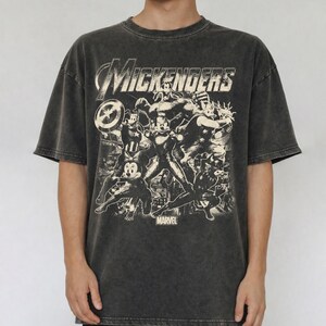 以下が含まれることがあります： チャコールグレーの酸洗い加工が施された半袖Tシャツ。スーパーヒーローの格好をした漫画キャラクターのグラフィックプリントが特徴です。上部には「MICKENGERS」の文字があり、グラフィックの下部には小さなフォントで「MARVEL」という言葉がプリントされています。