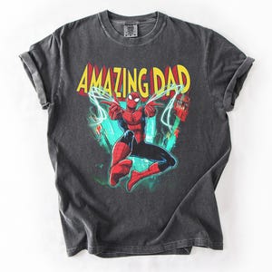 May include: Dark gray t-shirt featuring a colorful graphic of Spider-Man with the text "AMAZING DAD" in yellow and red. The superhero is in a dynamic pose with lightning bolts. The shirt is made of a soft material.