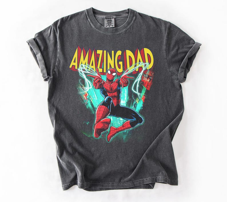 May include: Dark gray t-shirt featuring a colorful graphic of Spider-Man with the text "AMAZING DAD" in yellow and red. The superhero is in a dynamic pose with lightning bolts. The shirt is made of a soft material.