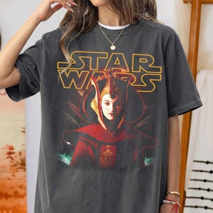 Puede incluir: Camiseta gr&aacute;fica gris oscuro de Star Wars con un retrato de un personaje con atuendo rojo y dorado. El logotipo de Star Wars es amarillo. La camiseta es de manga corta y cuello redondo.