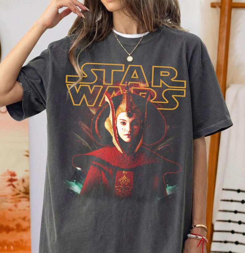 Puede incluir: Camiseta gr&aacute;fica gris oscuro de Star Wars con un retrato de un personaje con atuendo rojo y dorado. El logotipo de Star Wars es amarillo. La camiseta es de manga corta y cuello redondo.