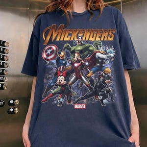 May include: A dark blue t-shirt featuring a graphic print titled MICKENGERS with cartoon characters dressed as superheroes. The design includes Mickey Mouse as Iron Man, Minnie Mouse as Black Widow, Goofy as Captain America, and others.