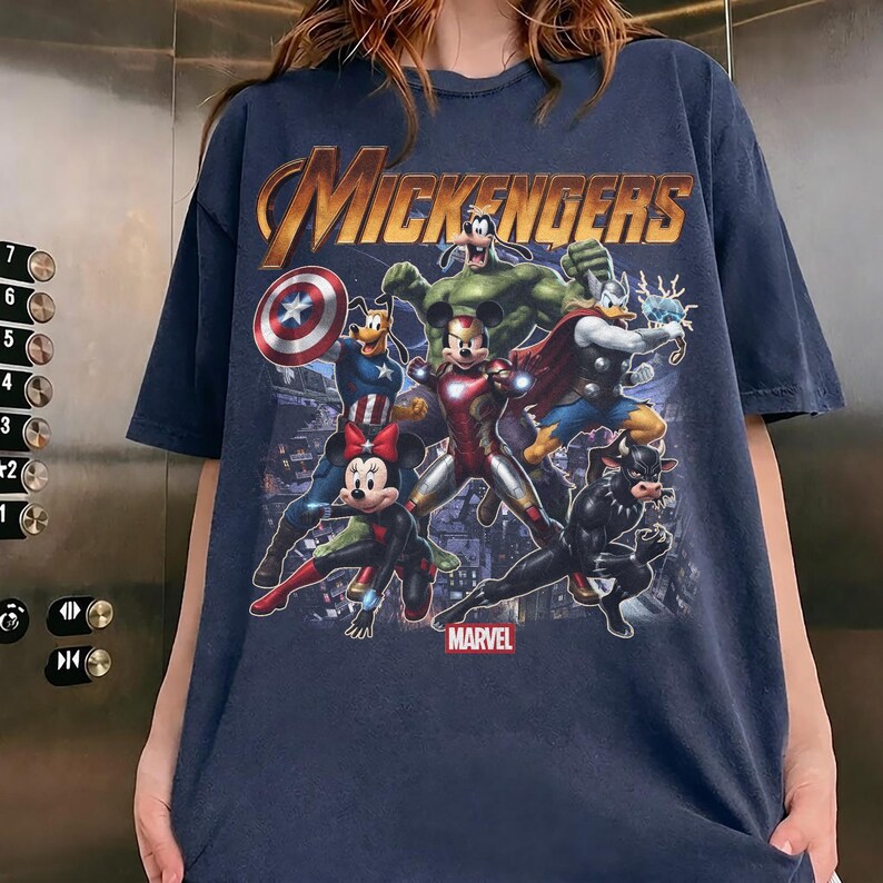 May include: A dark blue t-shirt featuring a graphic print titled MICKENGERS with cartoon characters dressed as superheroes. The design includes Mickey Mouse as Iron Man, Minnie Mouse as Black Widow, Goofy as Captain America, and others.
