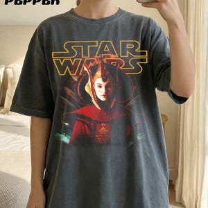 Puede incluir: Camiseta gris oscuro con un gr&aacute;fico de Star Wars. El texto amarillo dice "STAR WARS", y la imagen muestra un personaje con atuendo rojo y dorado. La palabra "PEPPER" est&aacute; impresa en la esquina superior izquierda.