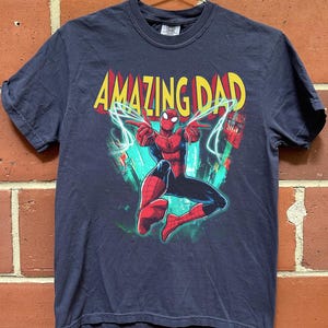 May include: Navy blue t-shirt featuring a graphic of Spider-Man with the text "AMAZING DAD" in yellow and red. The superhero is in a dynamic pose with lightning bolts. The shirt is hanging on a metal hanger against a brick wall.