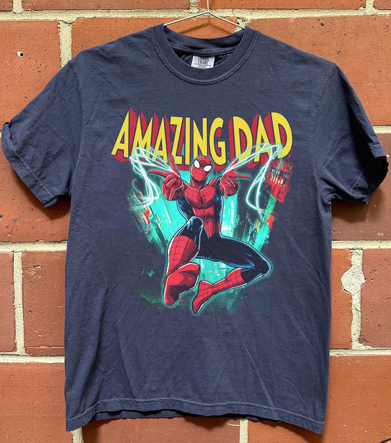 May include: Navy blue t-shirt featuring a graphic of Spider-Man with the text "AMAZING DAD" in yellow and red. The superhero is in a dynamic pose with lightning bolts. The shirt is hanging on a metal hanger against a brick wall.