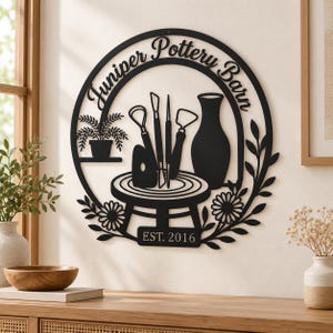 May include: A circular black metal wall sign featuring the text Juniper Pottery Barn and EST. 2016. The design includes silhouettes of pottery tools, a vase on a wheel, a potted plant on a shelf, and floral accents along the bottom edge.