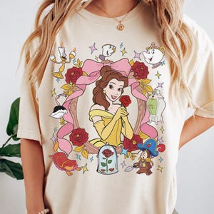 Puede incluir: Una camiseta de color crema con un estampado gráfico de Bella de La Bella y la Bestia. El diseño incluye un marco de cinta rosa, rosas rojas y varios personajes como Lumière, Din Don, la Sra. Potts, Chip y una rosa en una cúpula de cristal.