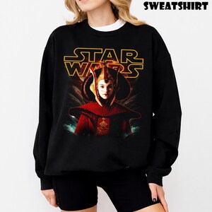 Puede incluir: Sudadera negra con un gr&aacute;fico de Star Wars, con el texto dorado "STAR WARS". La imagen muestra un personaje con un atuendo rojo y dorado. La palabra "SWEATSHIRT" est&aacute; impresa en la parte superior.