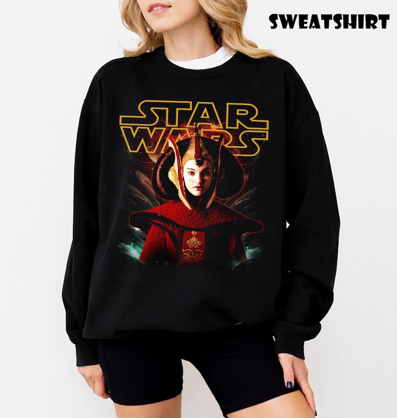Puede incluir: Sudadera negra con un gr&aacute;fico de Star Wars, con el texto dorado "STAR WARS". La imagen muestra un personaje con un atuendo rojo y dorado. La palabra "SWEATSHIRT" est&aacute; impresa en la parte superior.