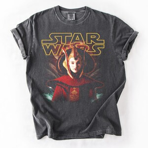 Puede incluir: Camiseta gris oscuro con el logo de Star Wars en dorado y un retrato de un personaje con atuendo rojo y dorado. La camiseta tiene un aspecto vintage y desgastado.