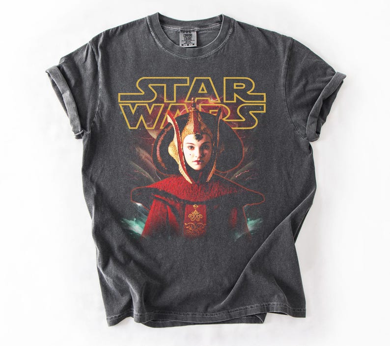 Puede incluir: Camiseta gris oscuro con el logo de Star Wars en dorado y un retrato de un personaje con atuendo rojo y dorado. La camiseta tiene un aspecto vintage y desgastado.