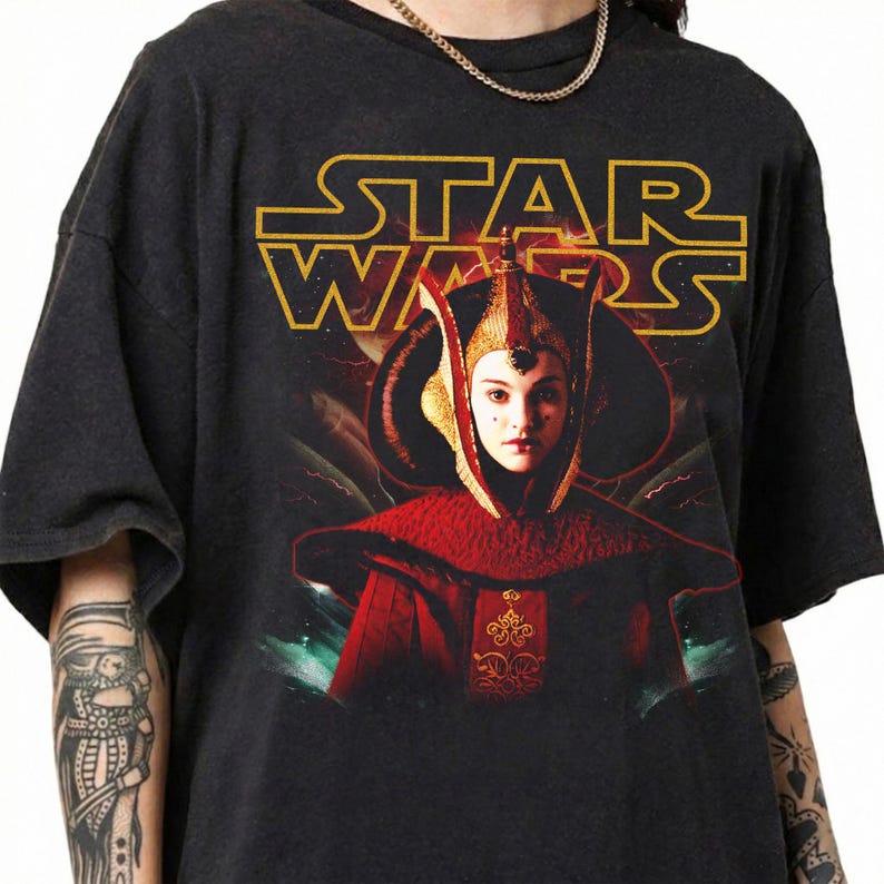 Puede incluir: Camiseta gris oscuro con el logo dorado de Star Wars y una imagen de un personaje con un traje rojo y dorado. El dise&ntilde;o gr&aacute;fico incluye un retrato del personaje y un fondo de monta&ntilde;as y nubes.