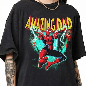 May include: Dark gray t-shirt featuring a colorful graphic of Spider-Man with the text "AMAZING DAD" in yellow and red. The superhero is in a dynamic pose, with web lines extending from his hands.
