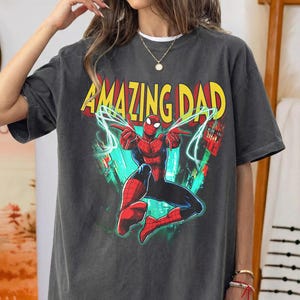 May include: Dark gray t-shirt featuring a graphic of Spider-Man with the words "AMAZING DAD" in yellow and red. The superhero is in a dynamic pose with blue energy lines. The shirt is a casual wear item.