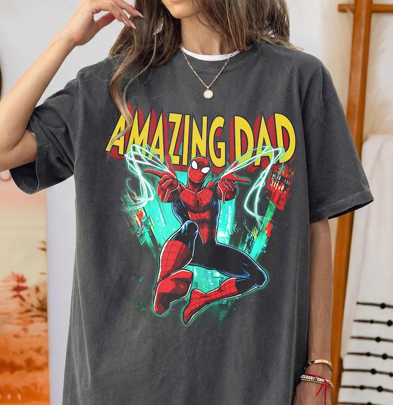 May include: Dark gray t-shirt featuring a graphic of Spider-Man with the words "AMAZING DAD" in yellow and red. The superhero is in a dynamic pose with blue energy lines. The shirt is a casual wear item.