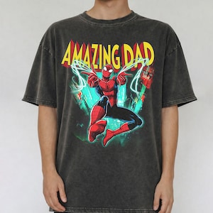 May include: Dark gray t-shirt featuring a graphic of Spider-Man with the text "AMAZING DAD" in yellow and red. The superhero is depicted in a dynamic pose with web-like energy effects. The shirt has a vintage, washed look.