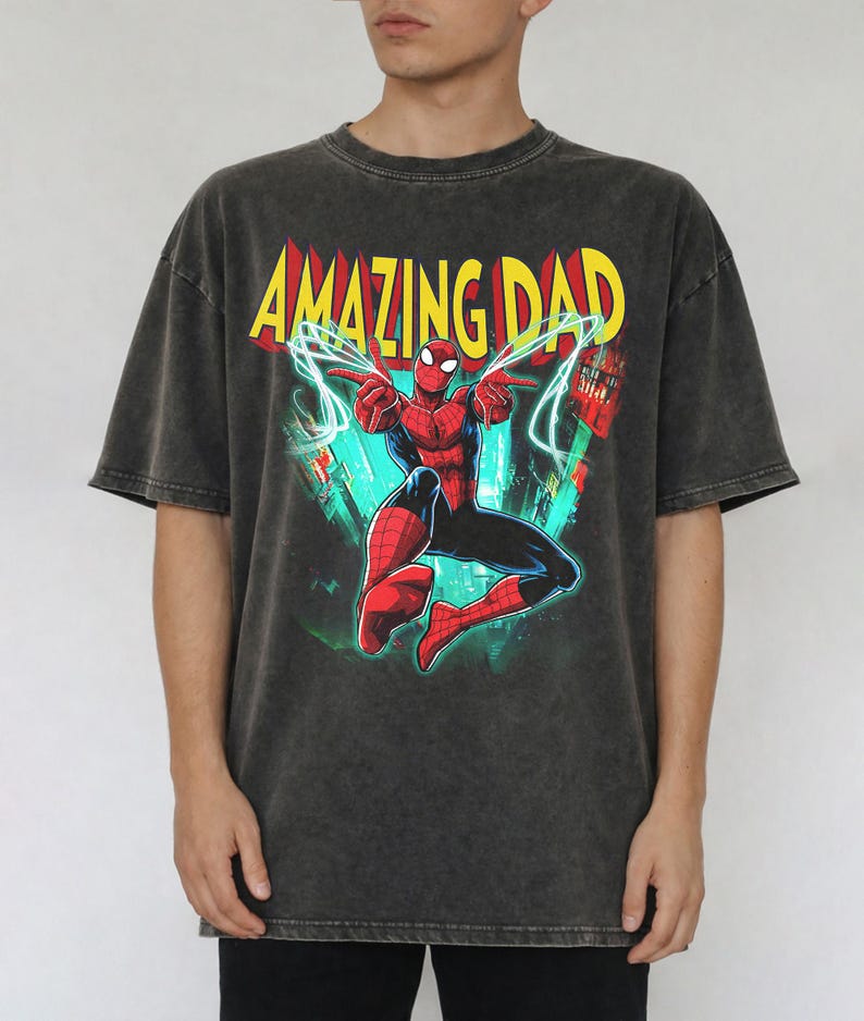 May include: Dark gray t-shirt featuring a graphic of Spider-Man with the text "AMAZING DAD" in yellow and red. The superhero is depicted in a dynamic pose with web-like energy effects. The shirt has a vintage, washed look.