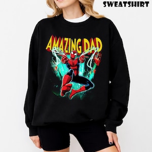 May include: Black sweatshirt featuring a colorful graphic of Spider-Man with the text "AMAZING DAD" in yellow and red. The word "SWEATSHIRT" is printed at the top. The design includes a cityscape background with green and blue accents.