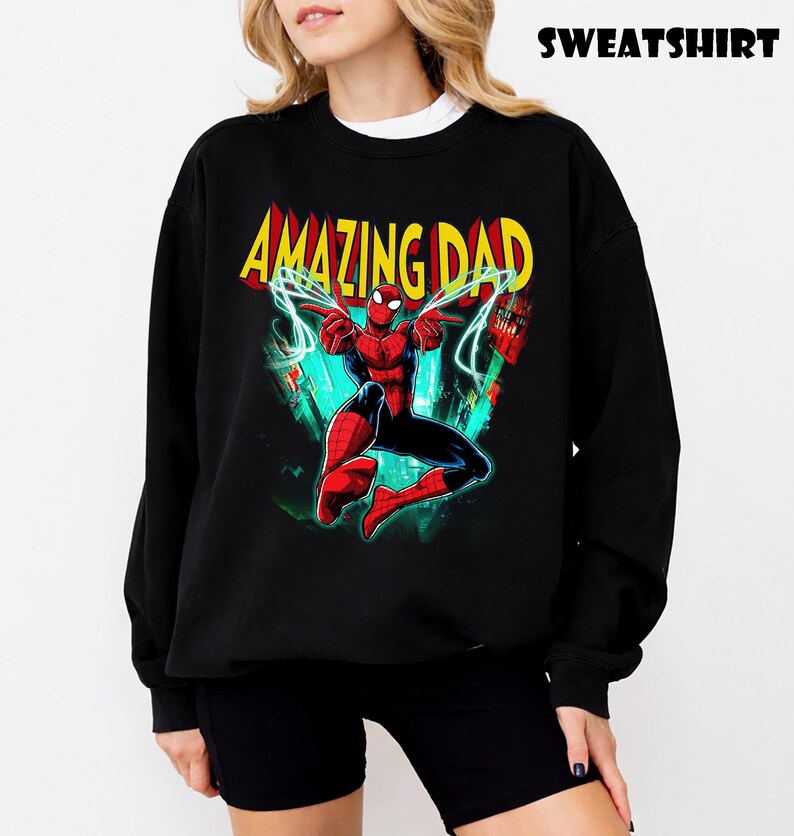 May include: Black sweatshirt featuring a colorful graphic of Spider-Man with the text "AMAZING DAD" in yellow and red. The word "SWEATSHIRT" is printed at the top. The design includes a cityscape background with green and blue accents.