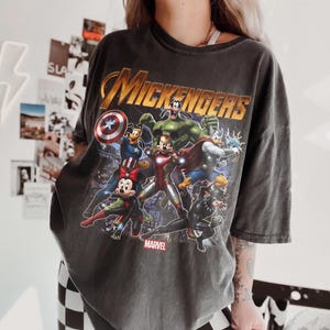 May include: A charcoal gray short-sleeve t-shirt featuring a graphic print of cartoon characters dressed as superheroes. The text "MICKENGERS" appears in gold at the top, with the "MARVEL" logo at the bottom. The design includes characters as Iron Man, Captain America, Thor, and Hulk.