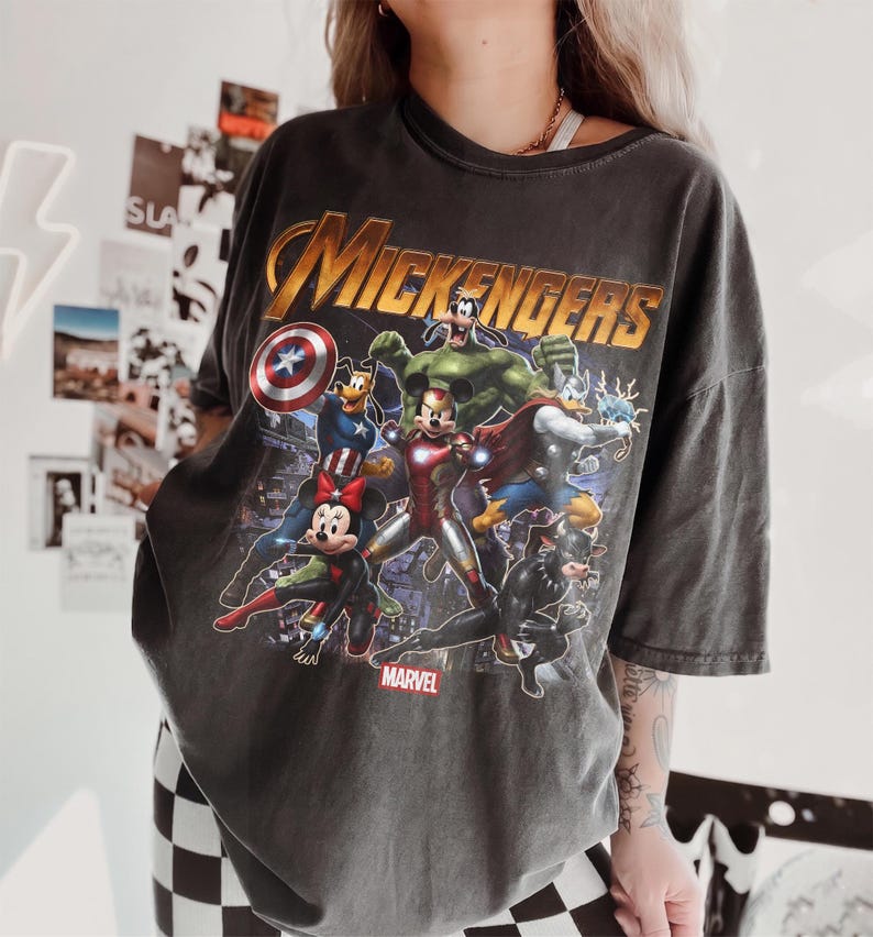 May include: A charcoal gray short-sleeve t-shirt featuring a graphic print of cartoon characters dressed as superheroes. The text "MICKENGERS" appears in gold at the top, with the "MARVEL" logo at the bottom. The design includes characters as Iron Man, Captain America, Thor, and Hulk.