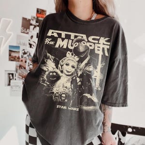 May include: A charcoal grey short-sleeve t-shirt featuring a distressed graphic print with the text "ATTACK OF THE MUPPET" and "STAR WARS" at the bottom. The design includes illustrations of Miss Piggy, Kermit the Frog, and other Muppet characters.