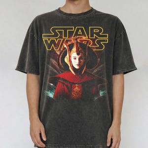 Puede incluir: Camiseta gris oscuro con el logo de Star Wars en amarillo y un gr&aacute;fico de un personaje con atuendo rojo y dorado. La camiseta tiene un aspecto vintage.