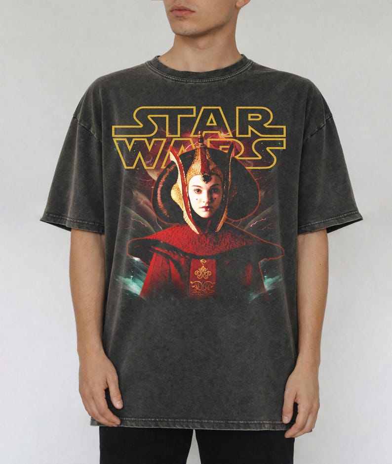 Puede incluir: Camiseta gris oscuro con el logo de Star Wars en amarillo y un gr&aacute;fico de un personaje con atuendo rojo y dorado. La camiseta tiene un aspecto vintage.