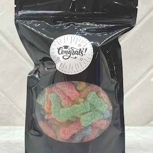 May include: A black, resealable bag containing colourful, sour gummy sweets. The sweets are shaped like small figures and are visible through a clear oval window. A white sticker with the word "Congrats!" and a graduation cap is affixed to the bag.