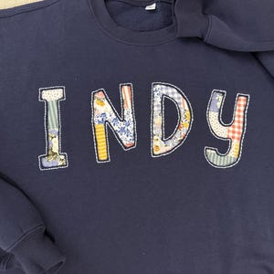 May include: A navy blue crewneck sweatshirt featuring the word INDY appliqued across the chest. Each letter is crafted from a different patterned fabric, including floral, gingham, and striped prints, all outlined with white decorative stitching.