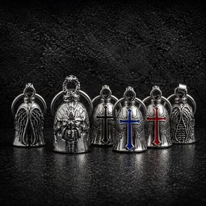 May include: A collection of six silver-toned metal bell pendants featuring various designs including angel wings, a detailed face, and crosses with blue and red enamel accents. Each bell is equipped with a circular ring at the top for attachment.